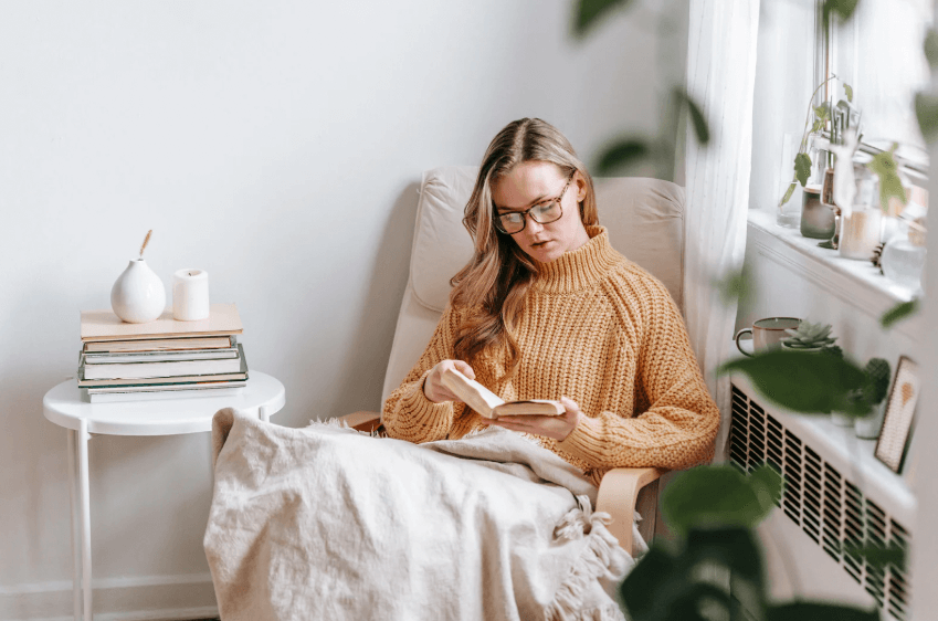 A photo of a woman in a yellow turtleneck sitting on a white couch with a book in her hand, while she is reading the book surrounded by plants and other books.