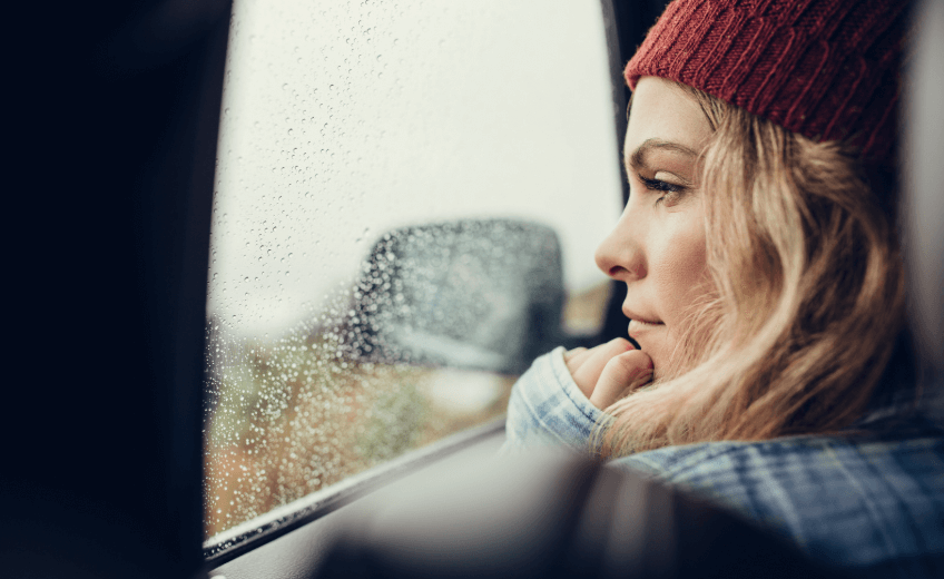 A photo of a woman in a blue flannel shirt and a red beanie in a car, while looking out the rainy window.