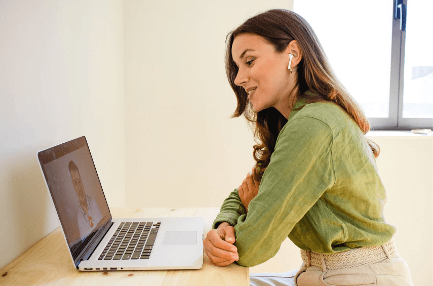 A photo of a woman in a green long-sleeve shirt smiling at a computer screen, while having a conversation with someone on the computer.