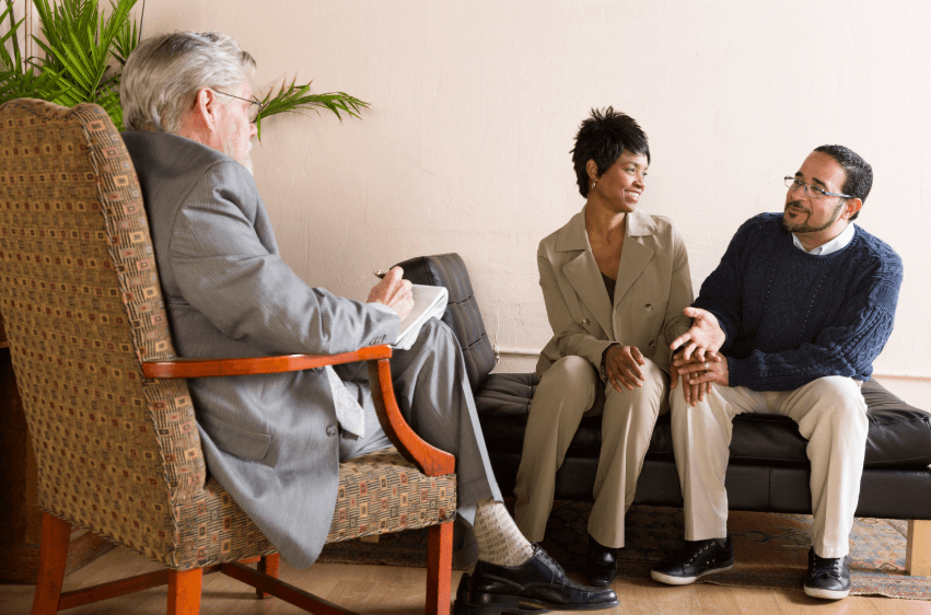 A photo of a couple sitting on a couch in front of a man portraying a therapist with a notebook in his hand. The couple is smiling and talking to the therapist.