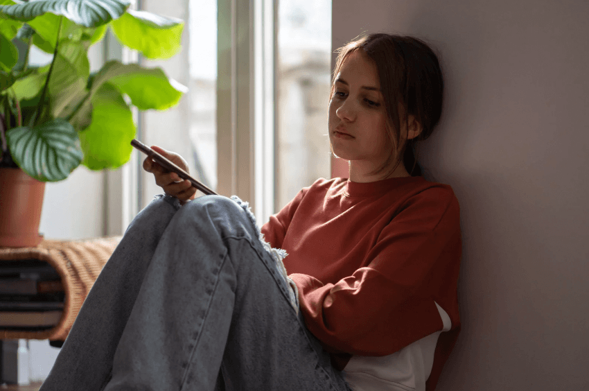 A photo of a young girl sitting with her back to a wall with a phone in her hand, with a slightly sad expression on her face.