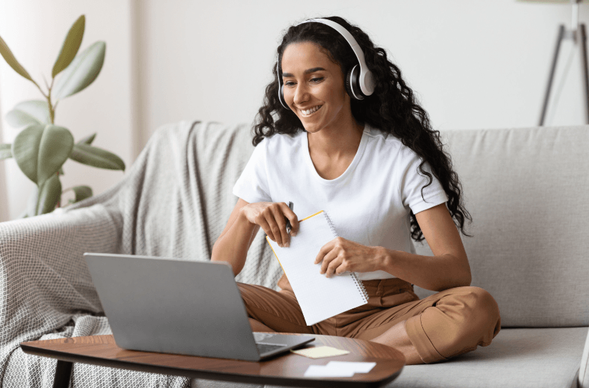 A photo of a woman with headphones on and sitting on a grey couch with a white notebook in hand, while smiling at a computer.