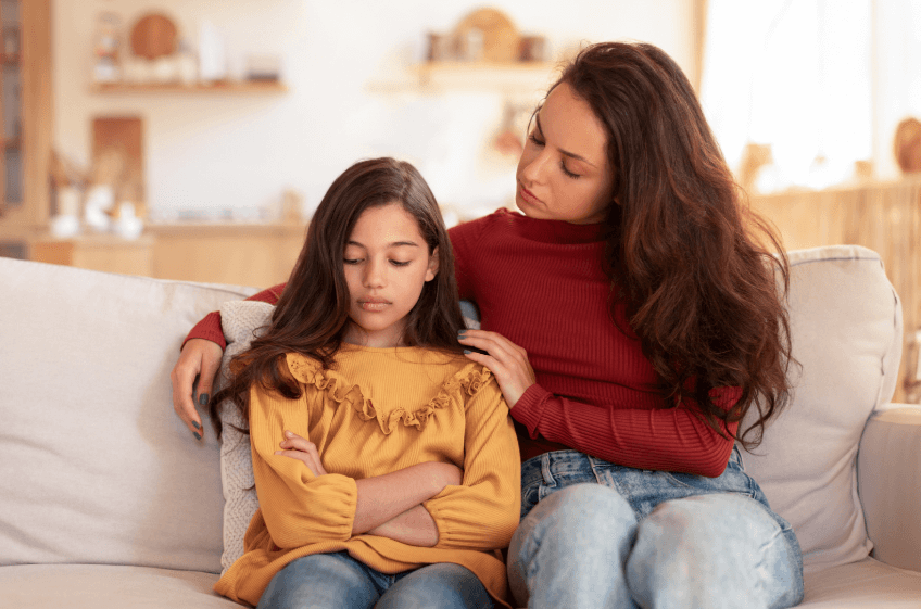 A photo of a mother and daughter sitting on a white couch, while the mother has her arms around her daughter and they have a serious expression on their faces.