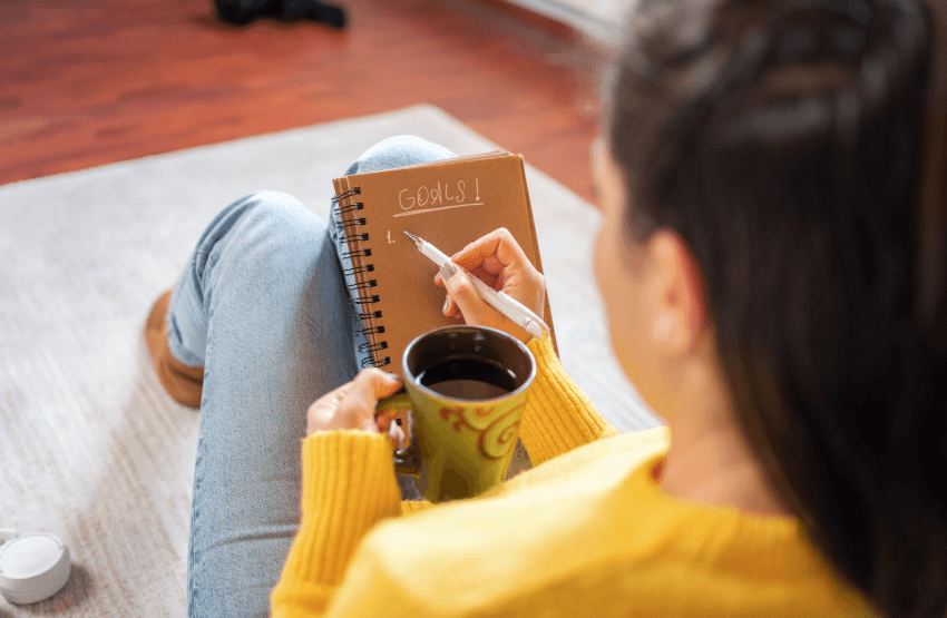 A photo of a woman in a yellow sweater and holding a coffee mug, while writing "Goals" on a piece of paper in a journal.