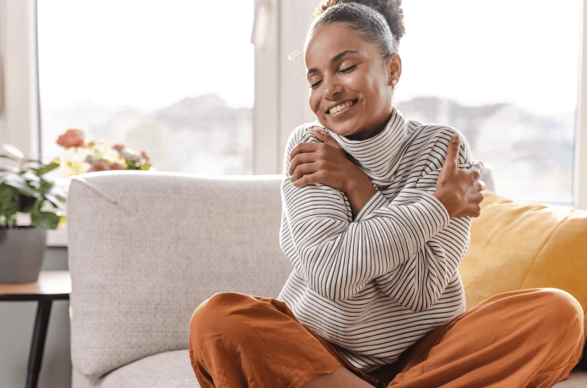 A photo of a woman sitting on a grey couch, while smiling and hugging herself.