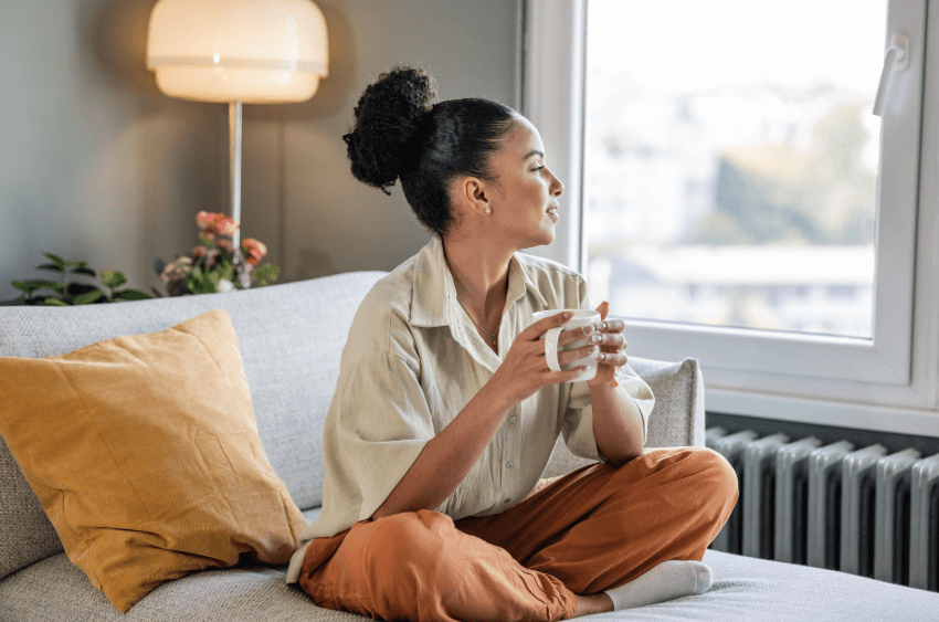 A photo of a woman sitting on a grey couch holding a white coffee mug, while looking out the window.