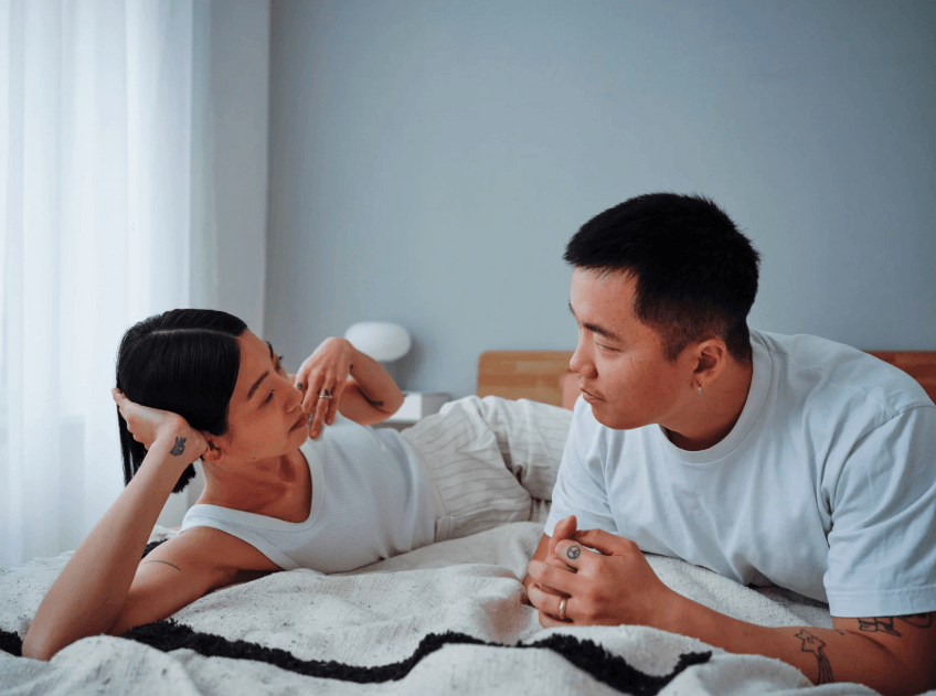 A photo of a couple laying on a bed looking at each other, while having an unemotional look on their faces.