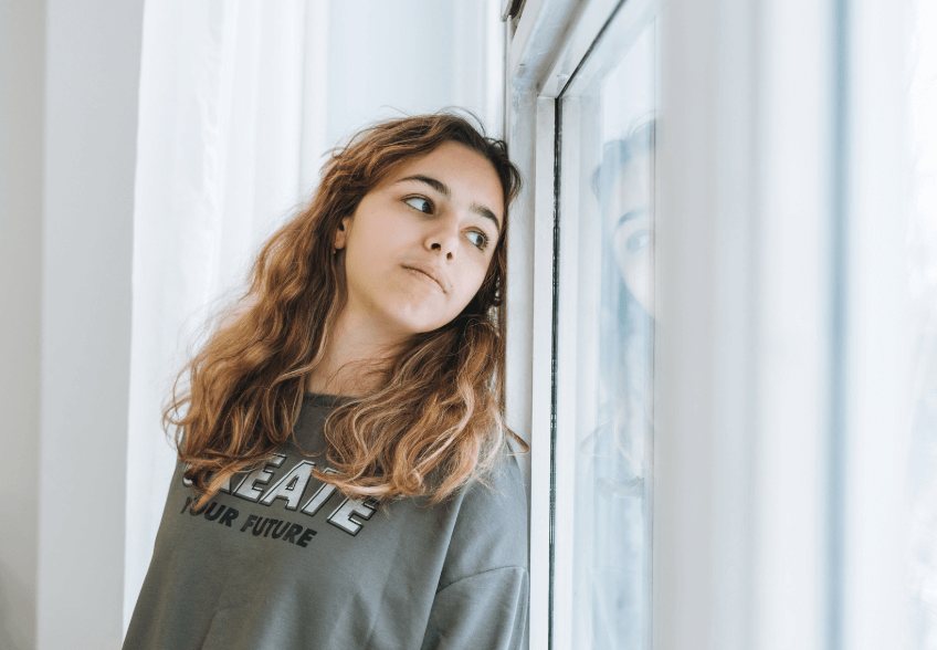 A photo of a young woman leaning up against a window, while looking out the window.