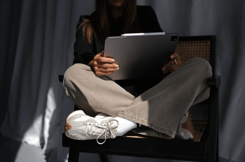 A photo of a woman sitting with her legs crossed on a chair, while holding a tablet.