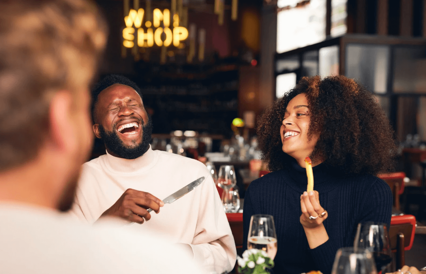 A photo of a couple eating dinner at a restaurant, where the man is laughing and holding a butter knife and the woman is smiling and holding a french fry.