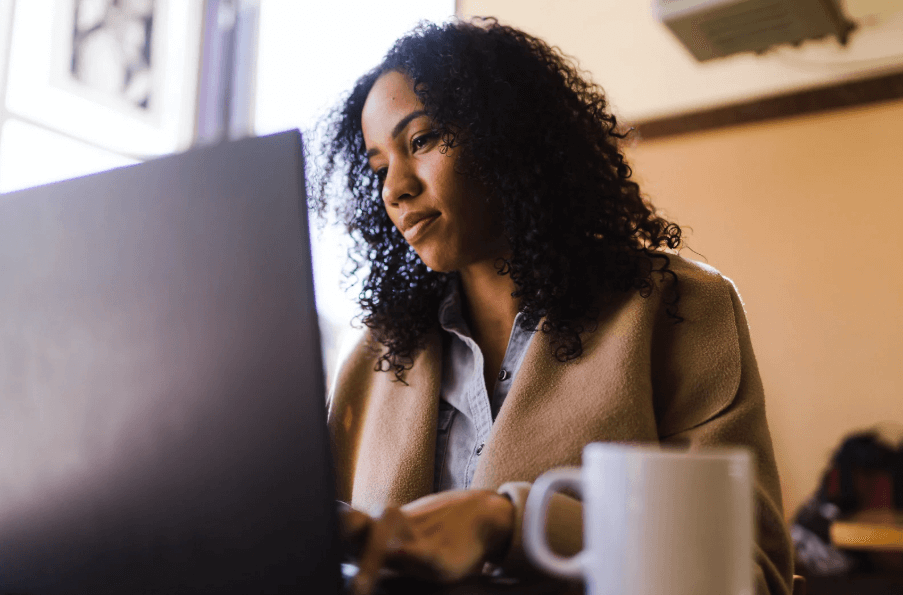 A photo of a woman in a beige jacket sitting in front of a computer screen with a white coffee mug next to the computer.