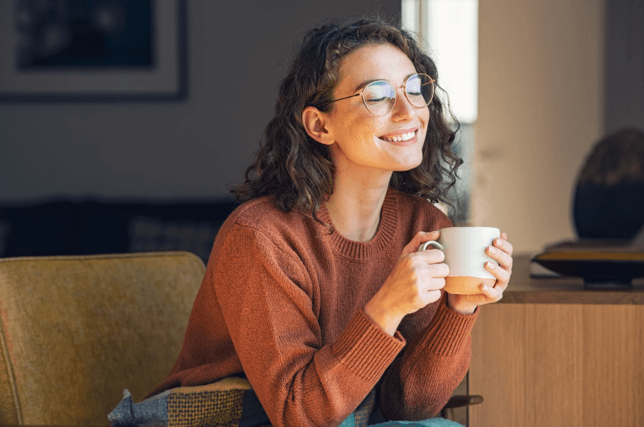 A photo of a woman in an orange sweater smiling, while holding a white coffee mug.