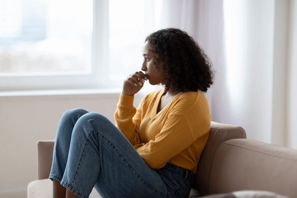 A photo of a woman in a yellow sweater, holding her hand to her face, while looking out the window to the left of her.